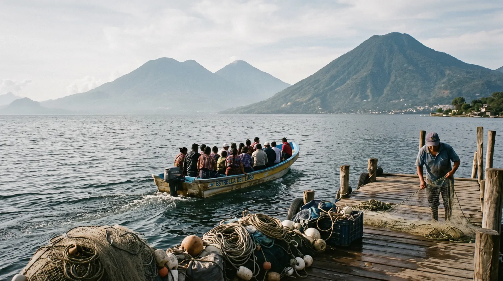 Lake Atitlan seen from the Panajachel dock at 09:00, volcanoes rising across the water, a wooden lancha passing with passengers seen from behind
